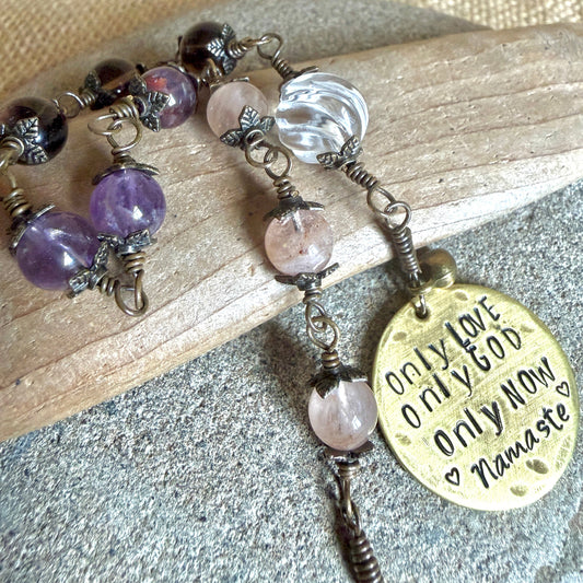 A beaded rosary with cacoxenite, morganite, and a crystal quartz bead, featuring a round brass pendant engraved with 'Only Love Only God Only Now Namaste'. The necklace is styled on a piece of wood over a gray stone surface.