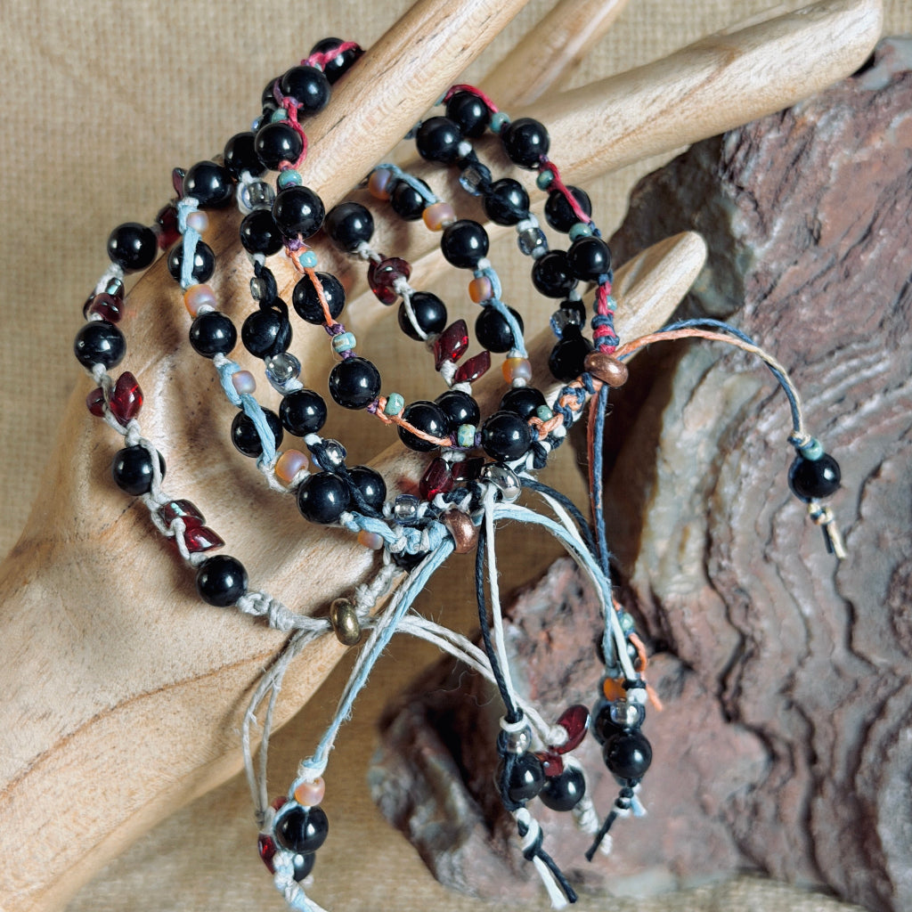 A set of handmade beaded bracelets with black round shungite beads, multicolor hemp cords, and small red, blue and orange accents, stacked on a wooden hand prop.