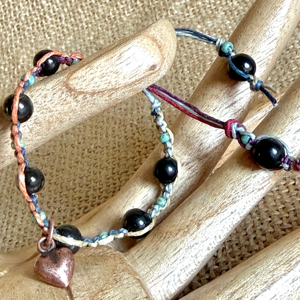 A handmade beaded bracelet with black shungite beads, multicolor hemp, and a small copper heart charm, displayed on a wooden mannequin hand against a burlap background.