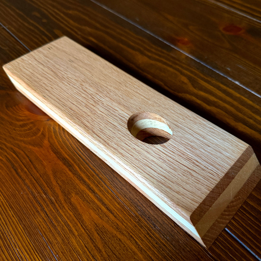 A rectangular wooden block with a circular cutout, featuring a light natural wood grain finish and a striped inlay inside the hole, shown on a dark wood tabletop.