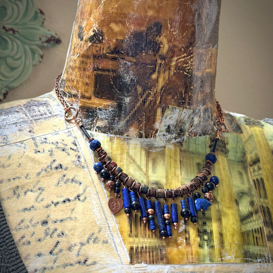 A beaded necklace displayed on a vintage-style collage mannequin bust. The necklace features copper-toned chain, antiqued copper beads, lapis lazuli beads, shungite beads, and a small dangling copper heart charm.