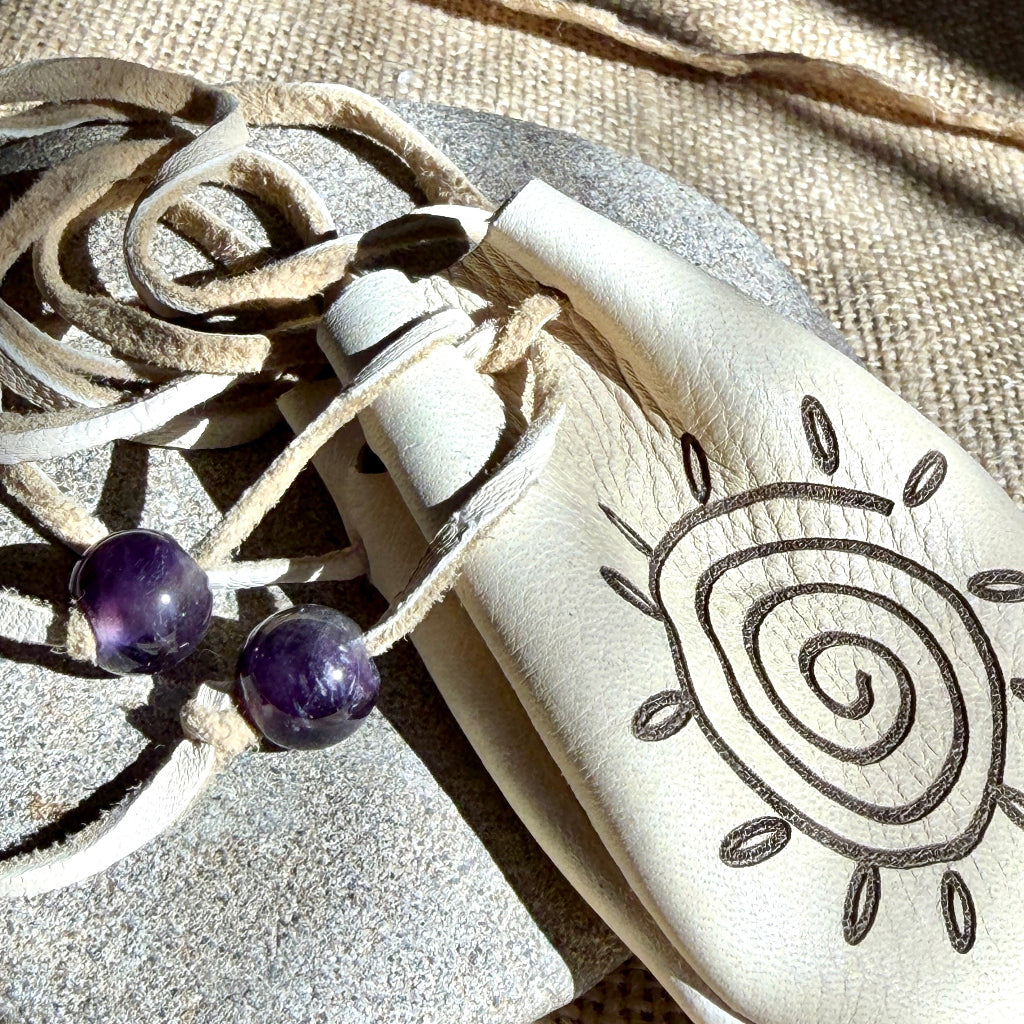A cream-colored leather pouch with a hand-drawn spiral sun design, featuring a drawstring closure with two purple amethyst beads, displayed on a textured burlap and stone background.