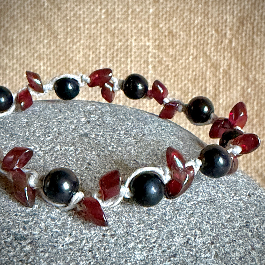 A beaded bracelet with glossy black round shungite beads and translucent dark red leaf-shaped beads, strung on natural cord and displayed on a gray stone against a tan fabric background.