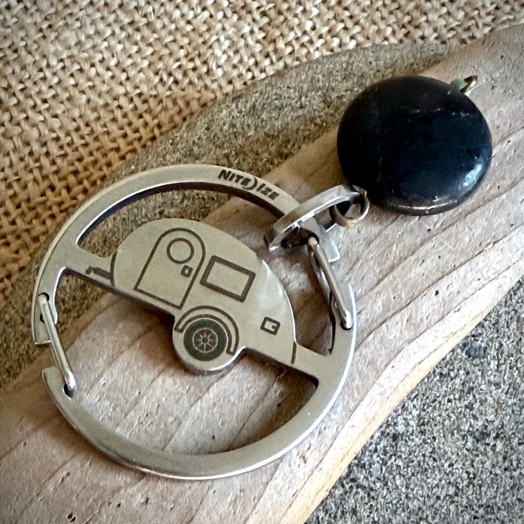 A silver-toned metal keychain featuring a circular design with a small camper trailer motif in the center, attached to a black shungite stone, photographed on a wooden surface with a woven textile background.