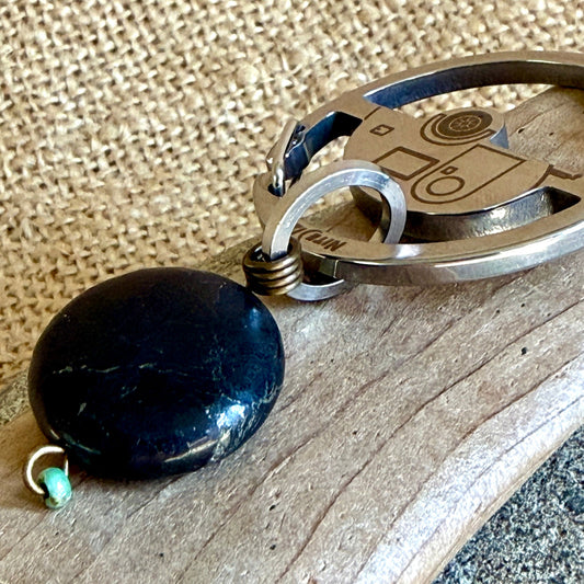 A close-up of a keychain with a polished shungite dangle, a silver metal keyring, and a small turquoise bead accent, resting on a wooden surface with a burlap background.