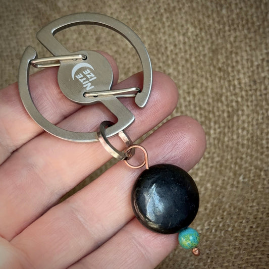 A close-up of a hand holding a metal keychain with a circular silver-tone ring, a round engraved center piece, and a dangling black oval stone charm with a small blue-green bead below.