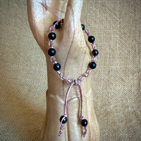 A beaded bracelet with black round shungite beads and pink woven hemp cord, displayed on a carved wooden hand form against a burlap background.