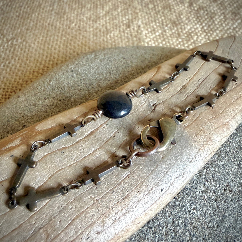 A handmade rosary-style bracelet with multiple small metallic crosses, a black shungite bead, and a bronze-toned clasp, displayed on a piece of weathered wood over a burlap fabric background.