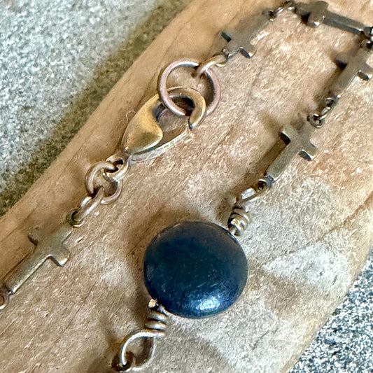 A close-up of a rosary-style bracelet with antique bronze-toned crosses and chain links, a round shungite coin bead, and a lobster clasp, resting on a textured tan surface.