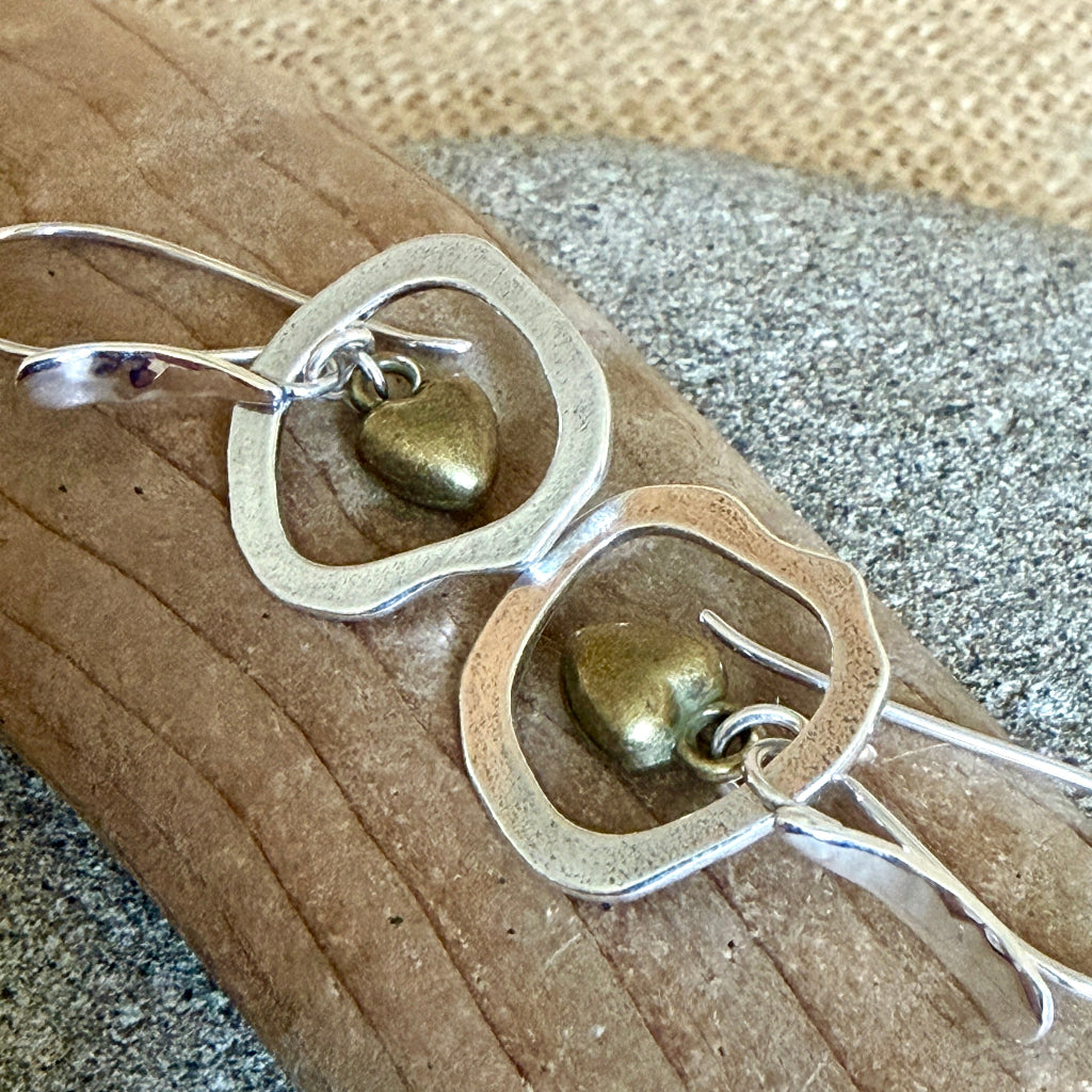 A pair of handmade dangle earrings with irregular silver-tone open circular frames and small antique bronze heart charms, displayed on a piece of driftwood over a burlap and stone background.