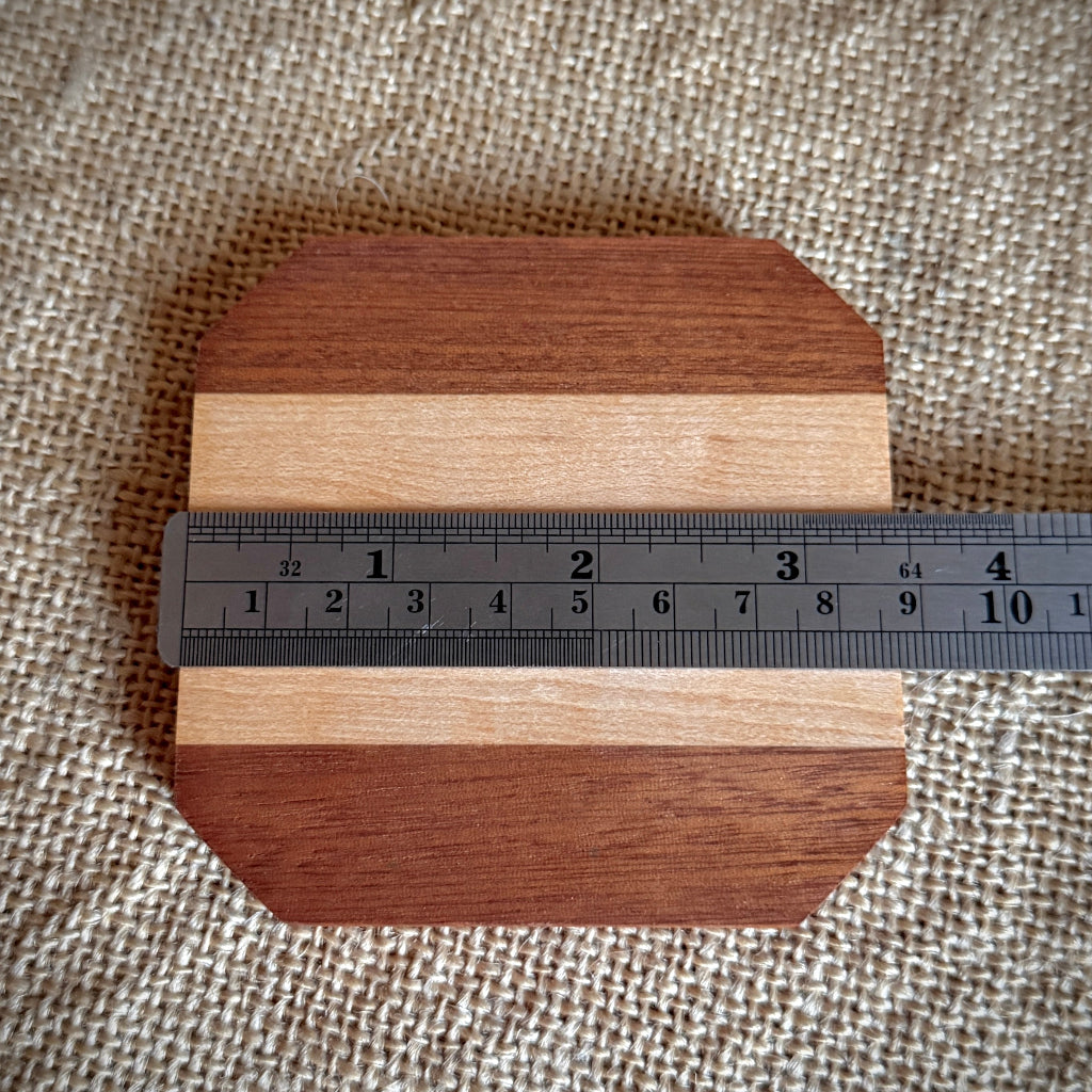 A small wooden board with rounded corners and alternating dark and light wood stripes, shown on a beige woven fabric background with a metal ruler across the middle for scale.
