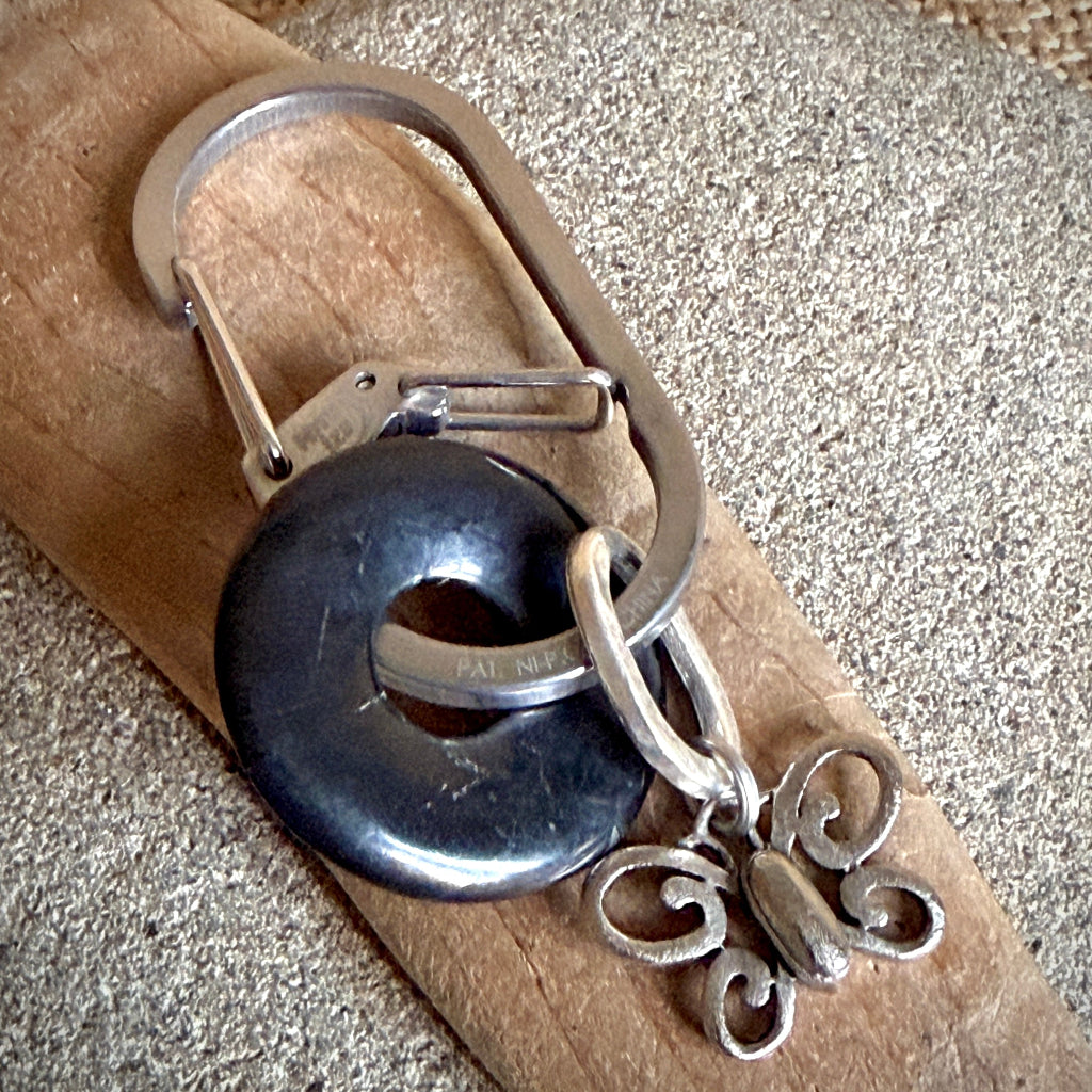 A handmade silver-toned keychain featuring a butterfly charm, a black shungite donut, and a carabiner, resting on a piece of wood over a textured gray surface.