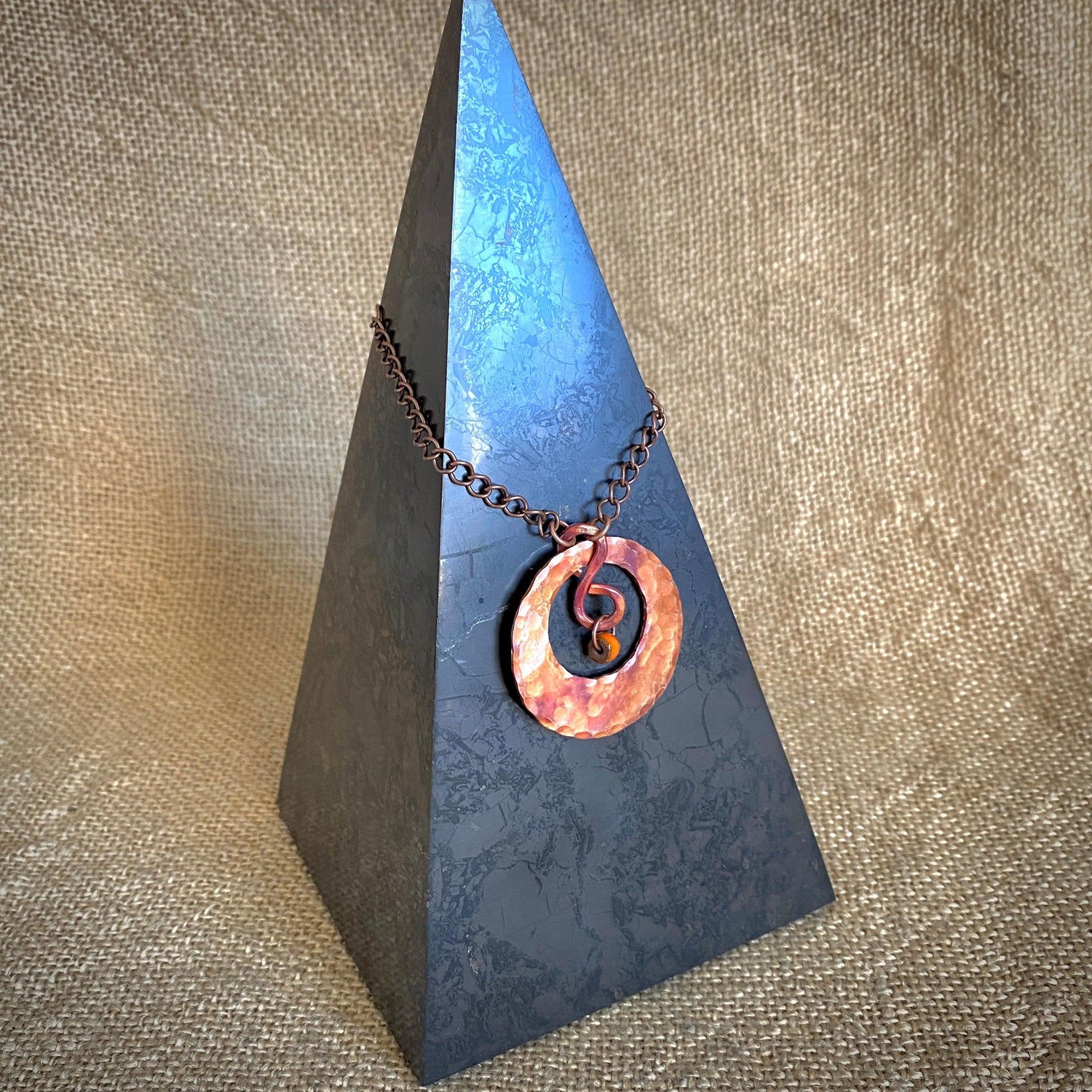 Hammered copper pendant necklace with a spiral design displayed on a dark grey marble pyramid stand.