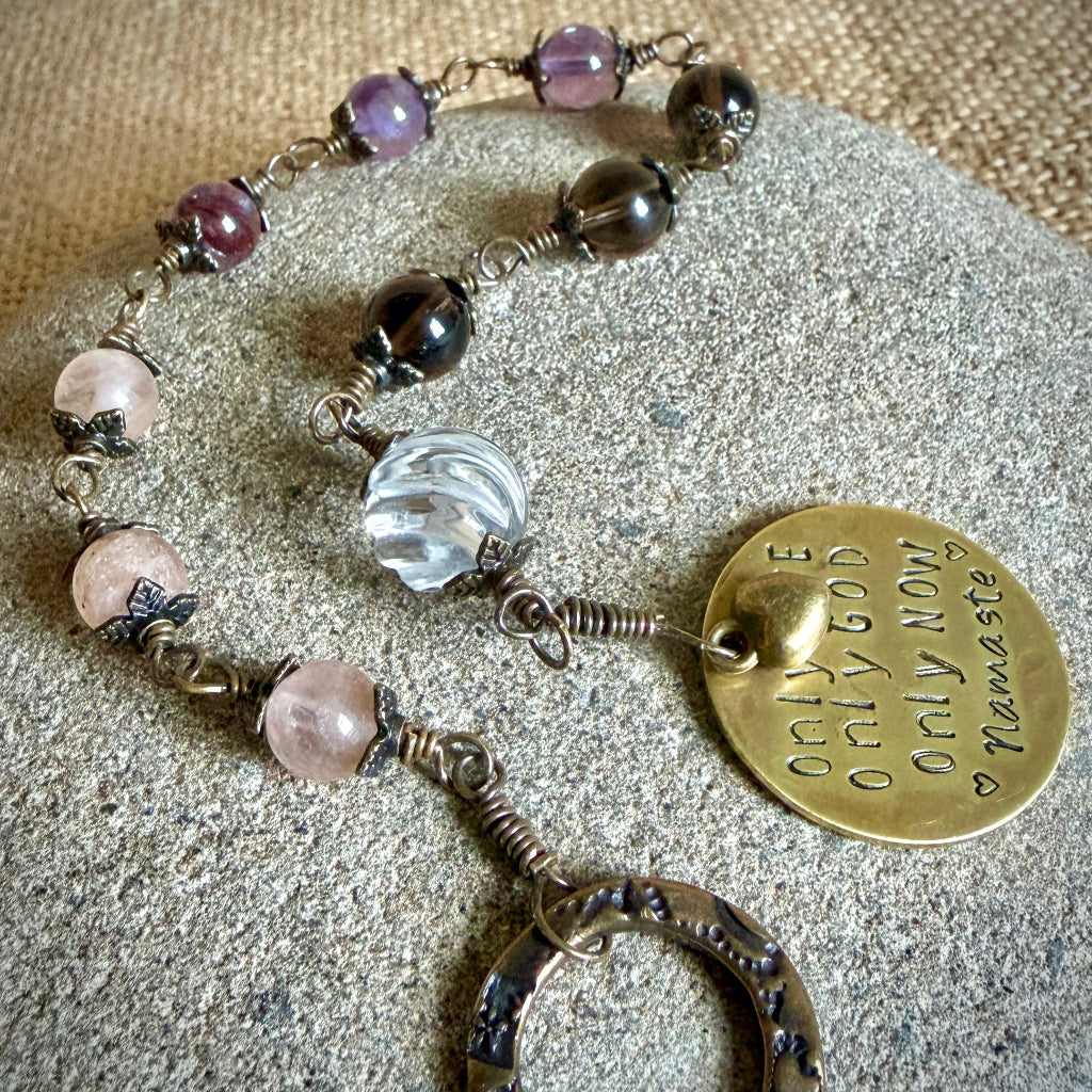 A handmade beaded rosary with a round brass disc charm engraved with 'Only Love Only God Only Now Namaste' and a large clear quartz bead, displayed on a textured gray stone.