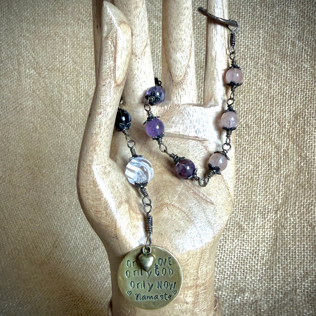 A beaded rosary with cacoxenite and morganite beads, a clear quartz bead, and a round engraved brass pendant reading 'ONE LOVE ONLY GOD ONLY NOW Namaste', displayed on a carved wooden hand against a beige fabric background.