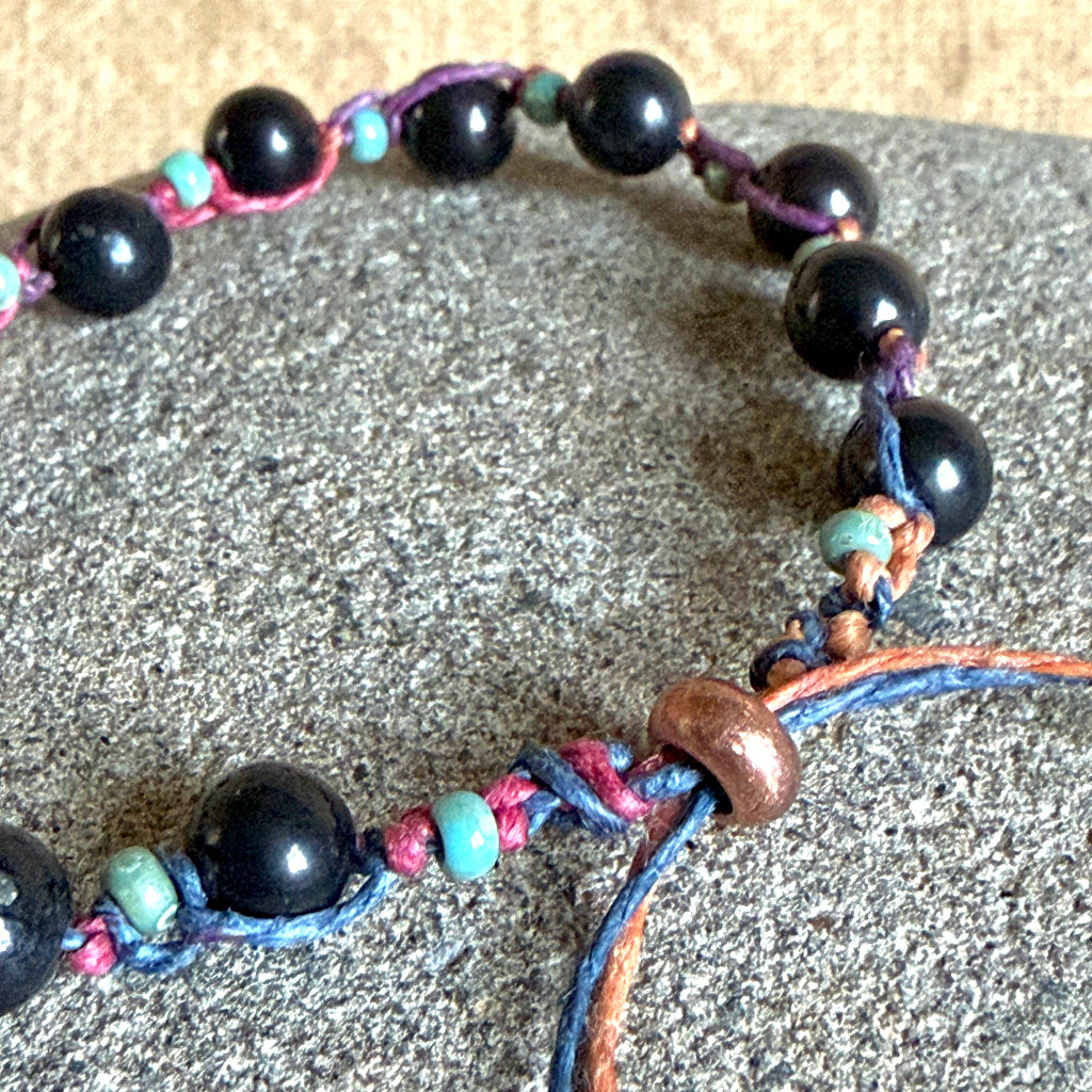 A beaded macrame bracelet with glossy black round shungite beads, turquoise spacer beads, and braided cords in purple, blue, orange, and pink, displayed on a textured gray stone surface.
