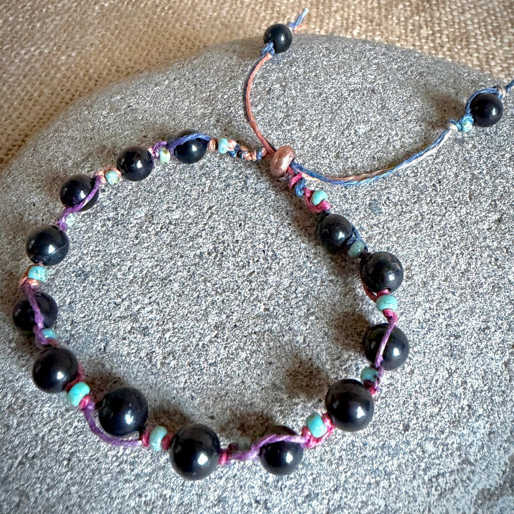 A beaded bracelet with large glossy black round shungite beads, small turquoise beads, and purple and blue cord, displayed on a round gray stone over a beige textured fabric background.