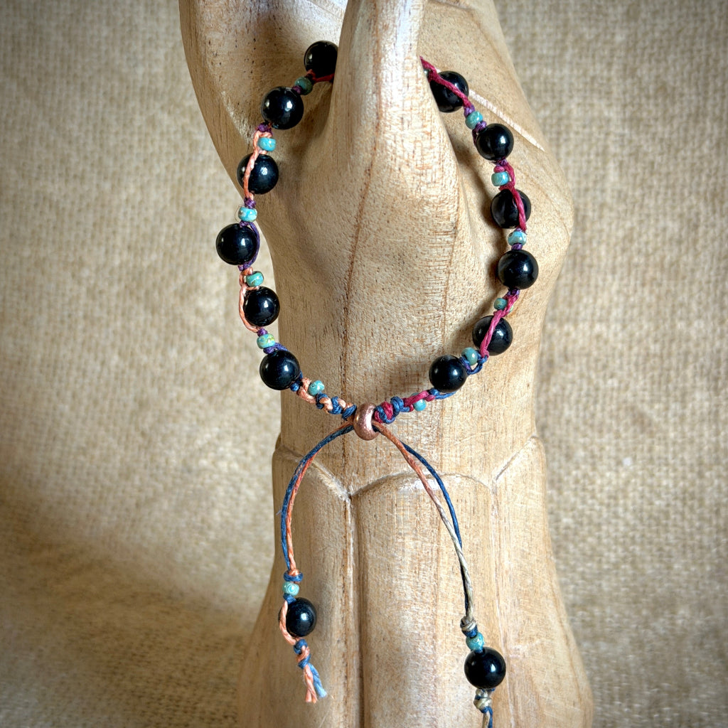 A beaded bracelet with glossy black round shungite beads and small turquoise accents, threaded on pink and blue cords, displayed on a carved wooden mannequin arm against a burlap background.