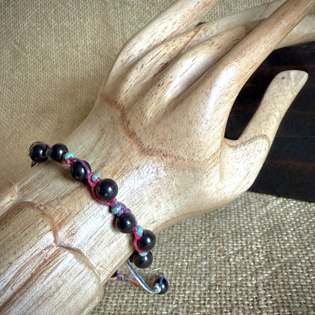 A beaded bracelet with glossy black round shungite beads and magenta cord, displayed on a carved wooden mannequin hand over a burlap background.