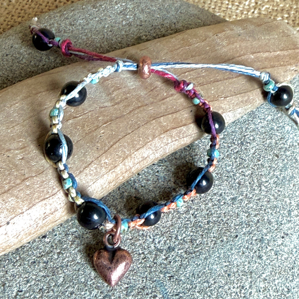 A handmade beaded bracelet with black shungite beads, blue and purple cord, and a small copper heart charm, displayed on a piece of wood over a gray textured surface.