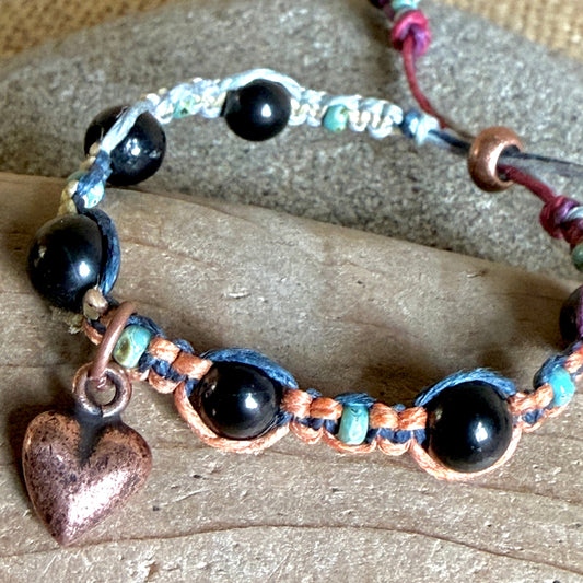 A handmade beaded bracelet featuring black shungite beads, woven copper and blue cord, turquoise accents, and a dangling copper heart charm, displayed on a piece of wood.