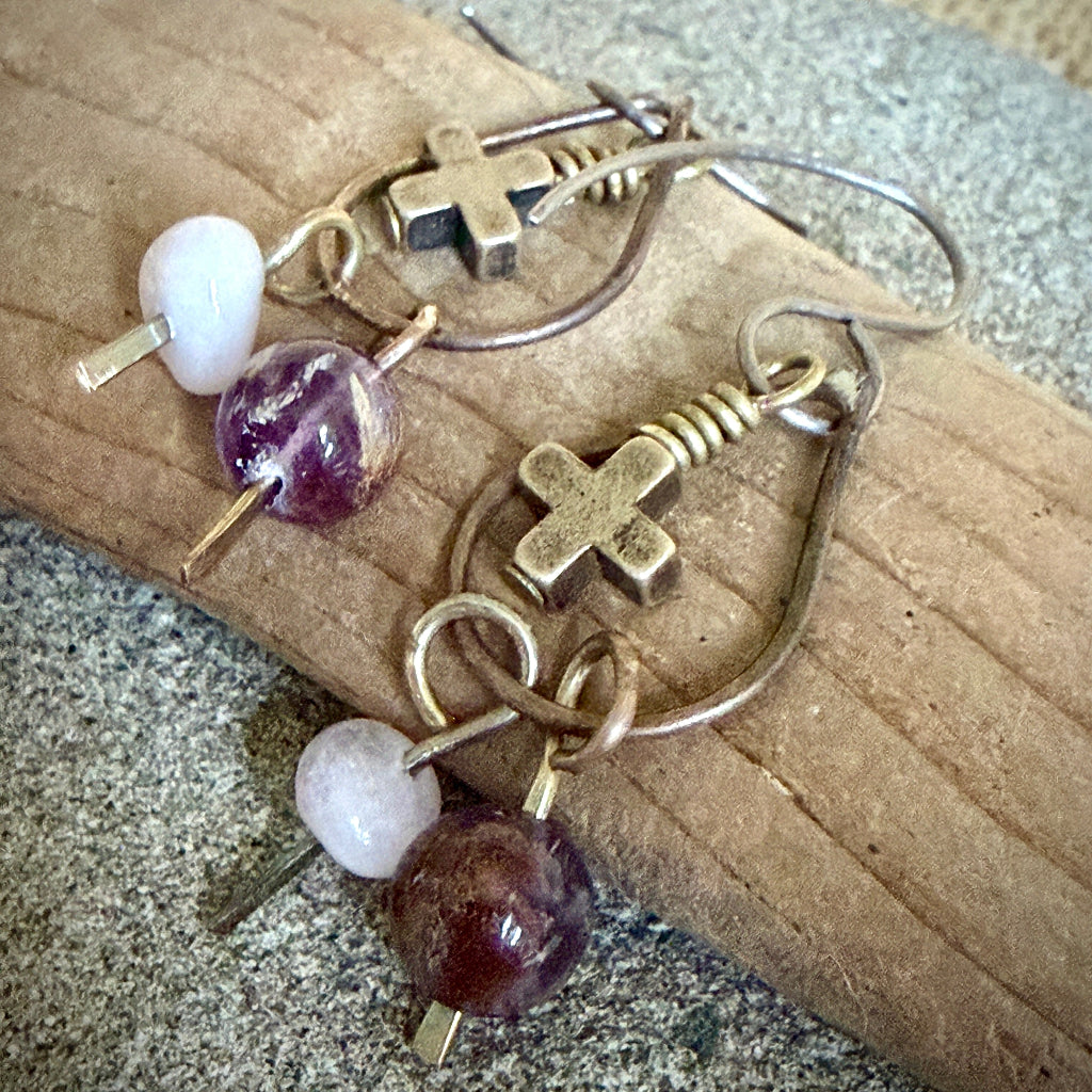 A pair of handmade dangle earrings with antique brass metal hooks, small cross charms, coiled wire details, a morganite bead, and a larger cacoxenite bead, displayed on a piece of wood over a stone surface.