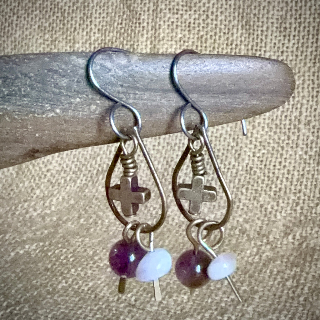 A pair of handmade dangle earrings with silver-toned metal hooks, brass-toned teardrop charms with crosses, and dangling beads in purple and white, displayed on a piece of wood against a burlap background.