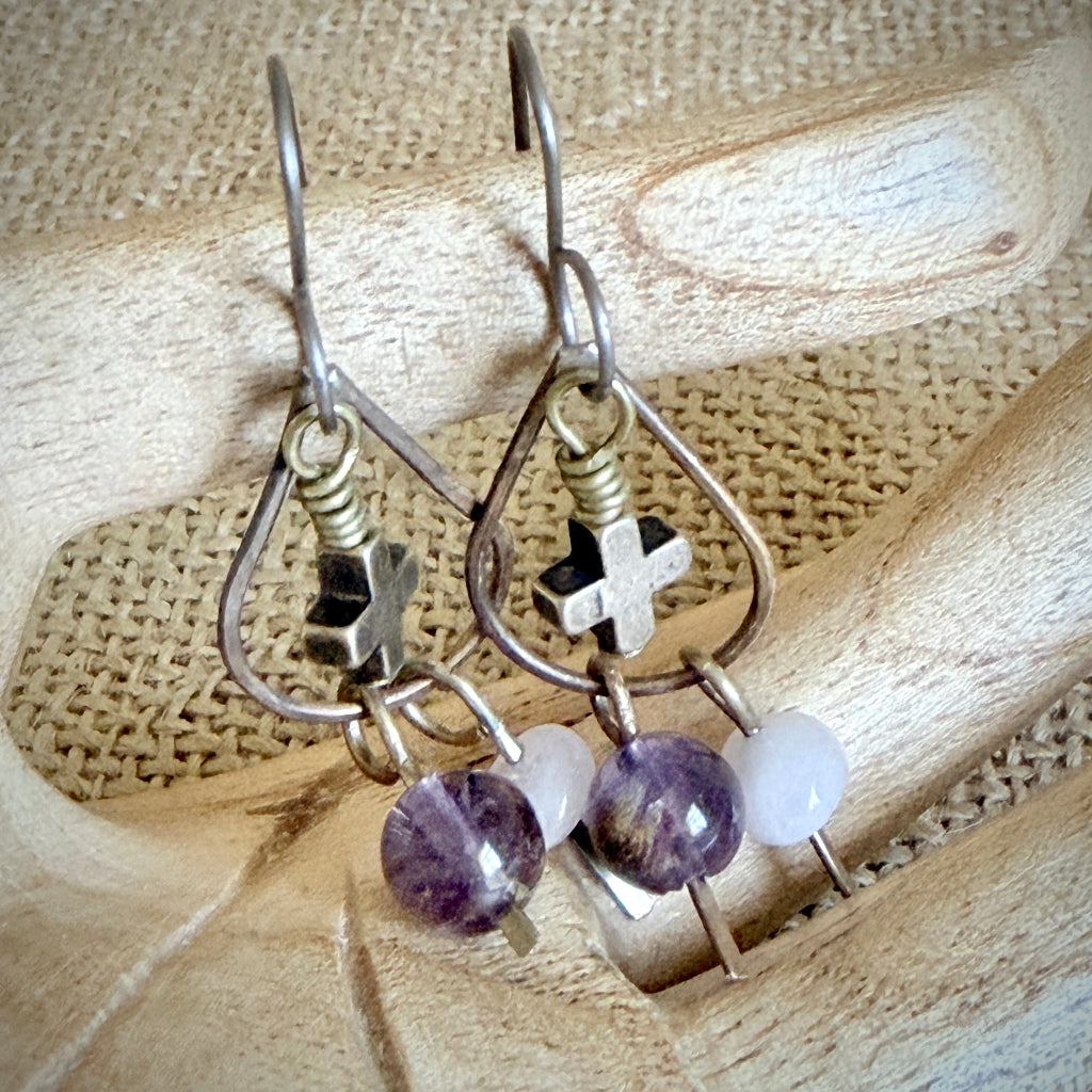 A pair of handmade dangle earrings displayed on a wooden hand. The earrings feature brass-toned metal hooks and frames with a small cross charm, a morganite bead, and a larger cacoxenite bead at the bottom.