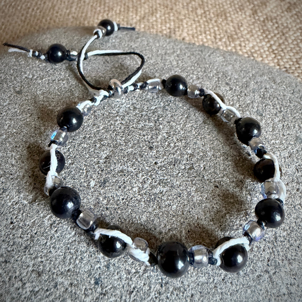 A beaded bracelet with black shungite beads, smaller clear glass beads, and black and white cord accents, displayed on a round gray stone surface.
