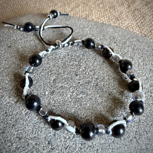 A beaded bracelet with black shungite beads, smaller clear glass beads, and black and white cord accents, displayed on a round gray stone surface.