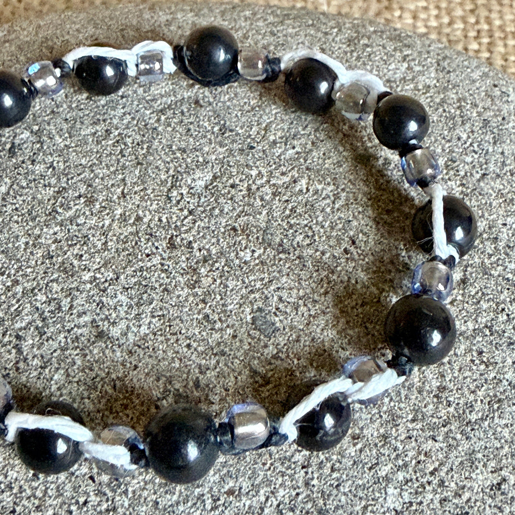 A beaded bracelet with glossy black round shungite beads, clear seed beads, and white and black woven cord, displayed on a textured gray stone surface.