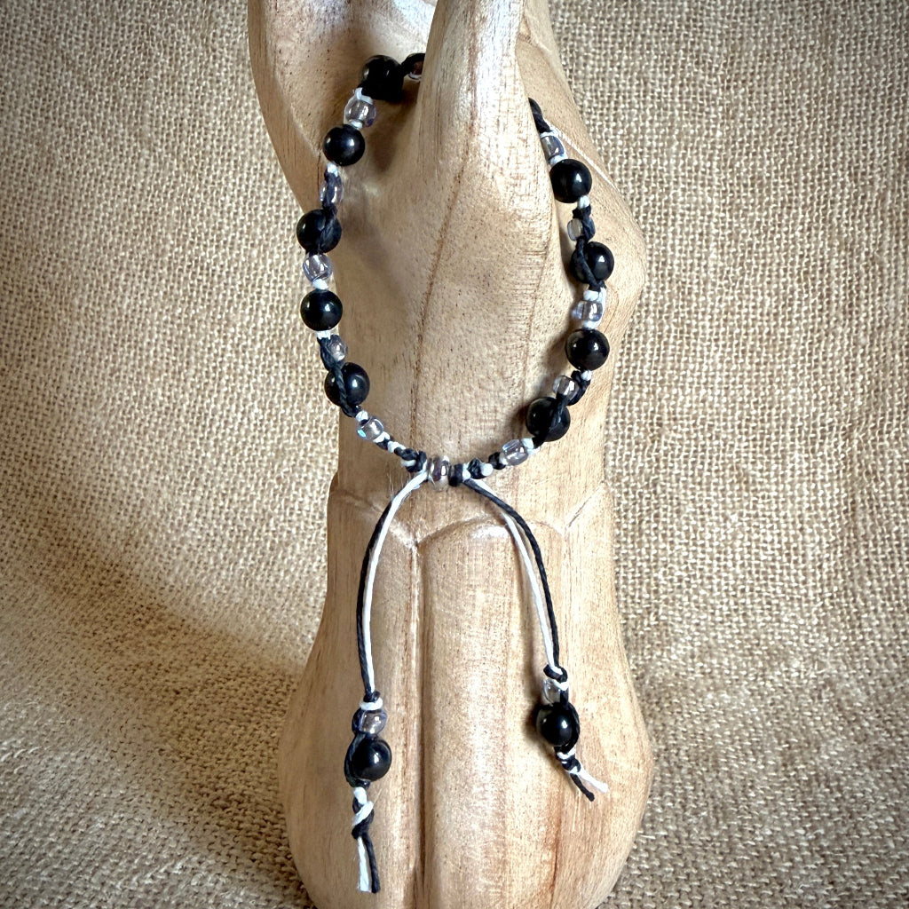 A beaded bracelet with black shungite beads, clear glass beads, and black and white accents, draped over a carved wooden display stand against a burlap background.