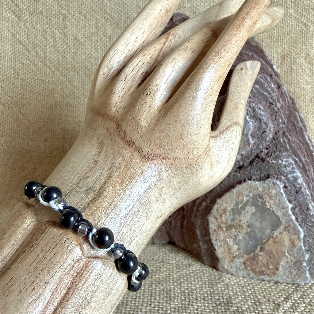 A beaded bracelet with glossy black shungite beads and small glass spacer beads, displayed on a carved wooden hand mannequin against a textured beige fabric background with a stone prop.
