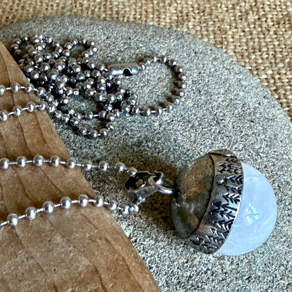 A silver ball-chain necklace with an acorn pendant resting on a stone and wood surface. The pendant features a textured silver cap and a white translucent quartz stone.