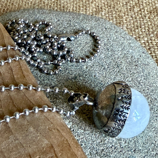 A silver ball-chain necklace with an acorn pendant resting on a stone and wood surface. The pendant features a textured silver cap and a white translucent quartz stone.