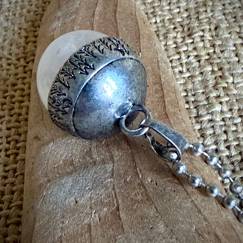 A close-up of a silver acorn pendant necklace with a white quartz insert, resting on a textured wooden surface over a woven burlap background.