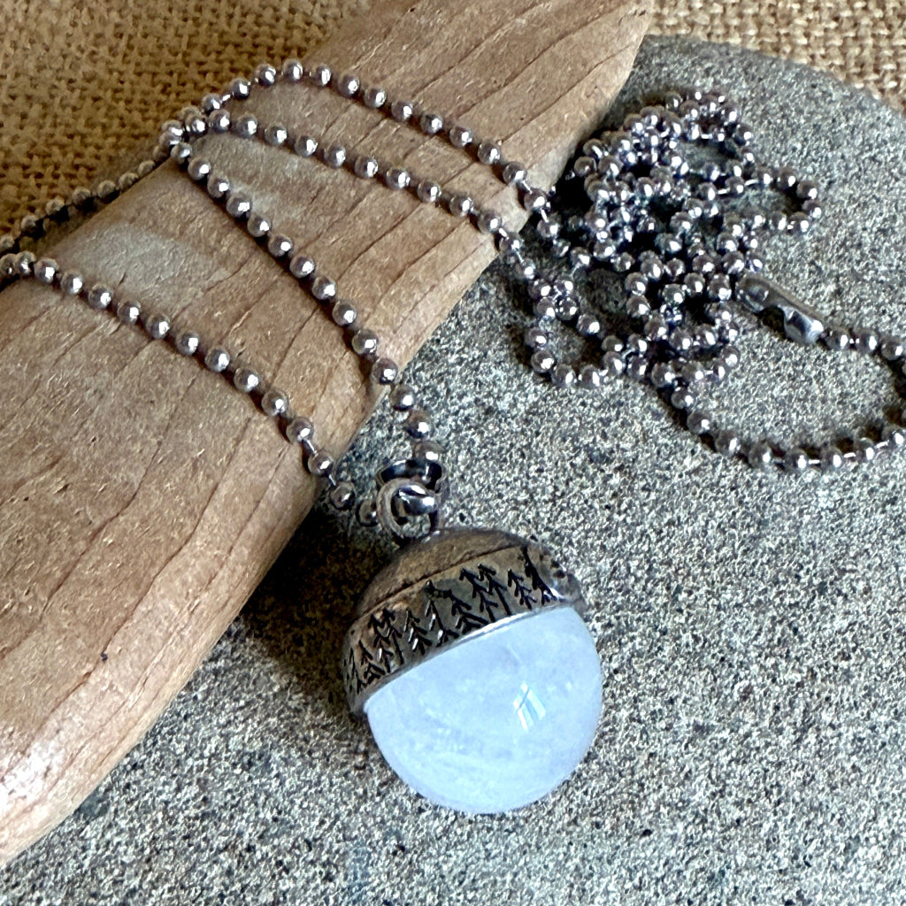 A silver-toned necklace with a round pendant featuring a textured silver top and a white quartz bottom, displayed on a piece of driftwood over a gray stone surface.