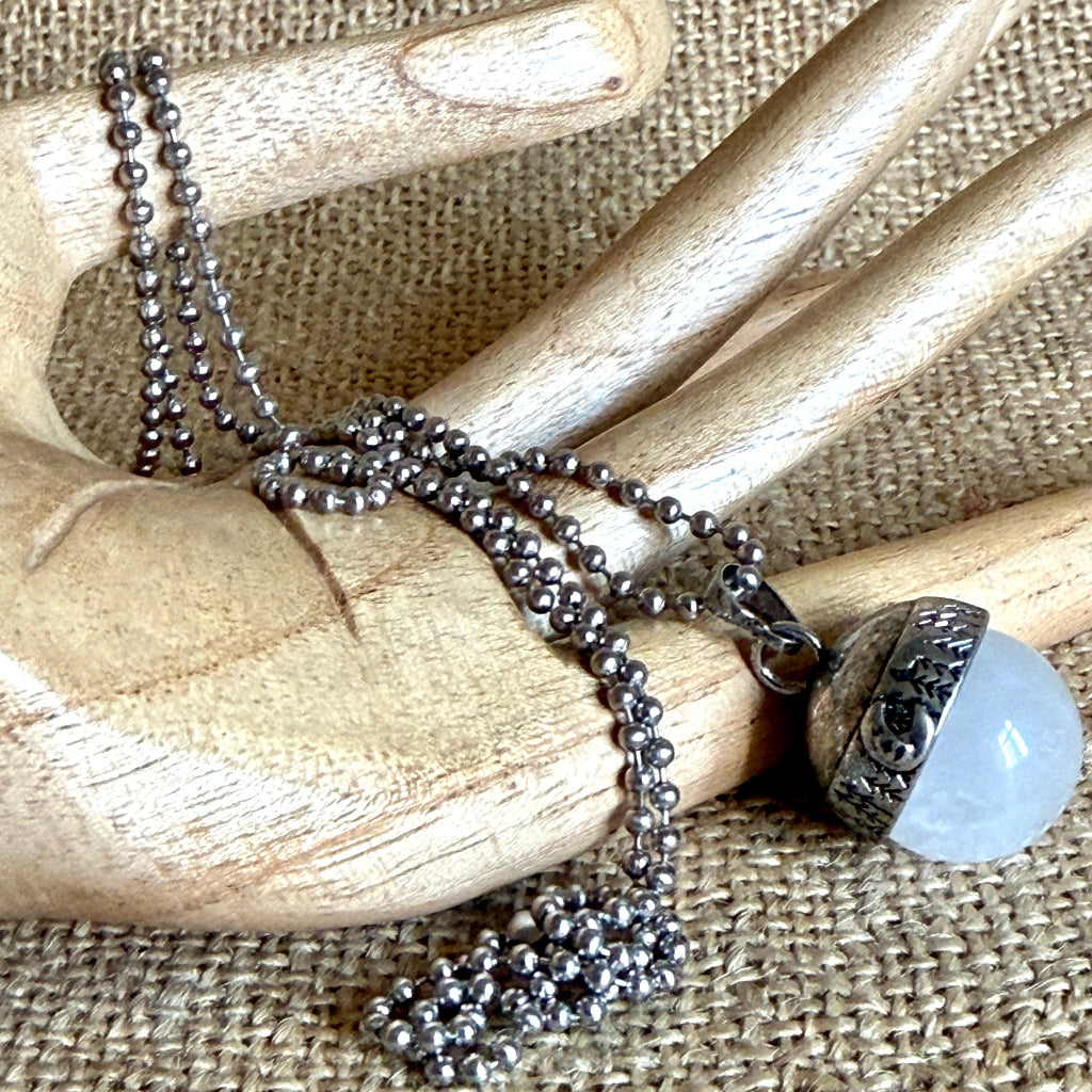 A silver-toned necklace with a large round white quartz pendant, displayed on a wooden hand against a burlap background.