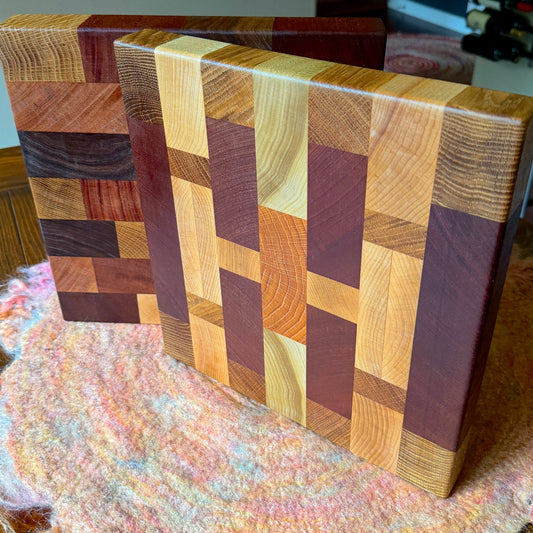 A close-up of a rectangular wooden cutting board with a multicolor geometric block pattern made from different wood tones, standing on a fuzzy pink and orange textured surface.
