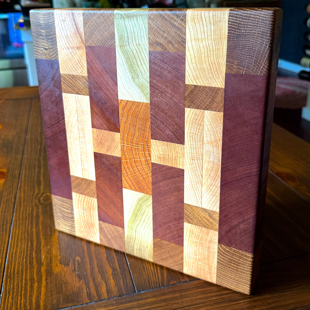 A square wooden cutting board with a geometric mosaic pattern of various wood grain panels in shades of brown, beige, and reddish-purple, standing on a dark wooden table.