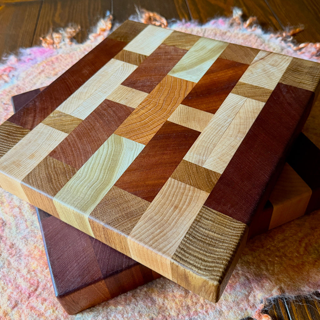 Two rectangular wooden cutting boards with a multicolor block pattern made from different shades of natural wood, resting on top of another wooden board on a pink fuzzy rug over a dark wood floor.