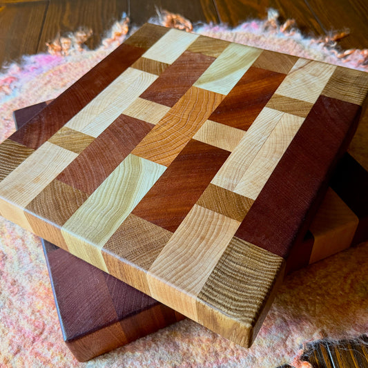 Two rectangular wooden cutting boards with a multicolor block pattern made from different shades of natural wood, resting on top of another wooden board on a pink fuzzy rug over a dark wood floor.