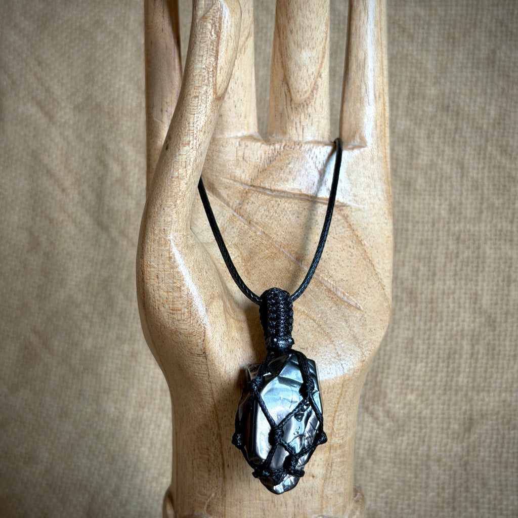 A black cord necklace with an elite shungite pendant displayed on a carved wooden hand against a neutral beige fabric background.