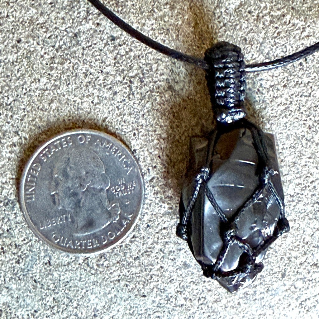 A black cord-wrapped elite shungite pendant necklace shown next to a US quarter coin for scale, on a textured gray surface.