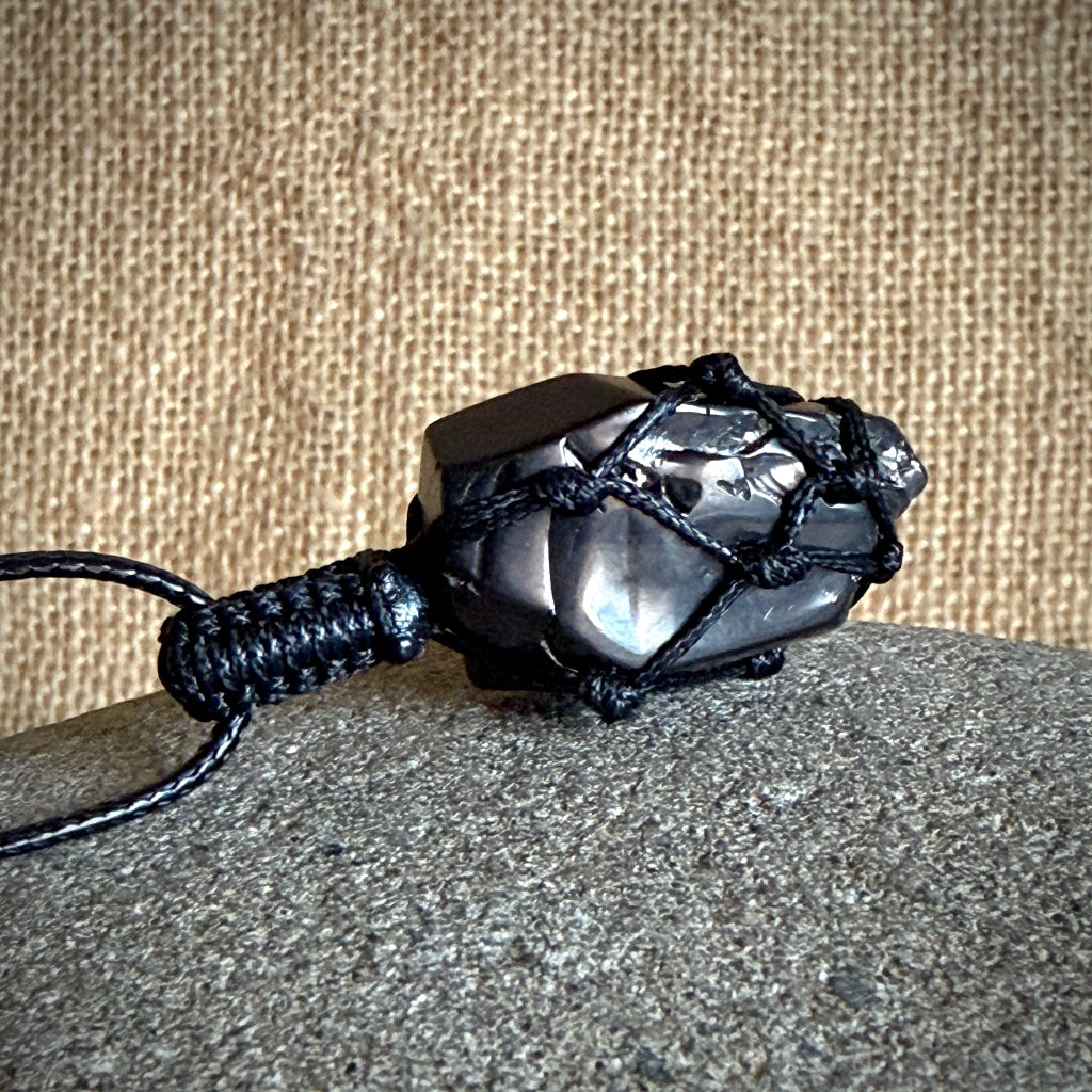 A dark gray to black elite shungite pendant wrapped in black cord, resting on a textured gray stone surface against a tan burlap background.