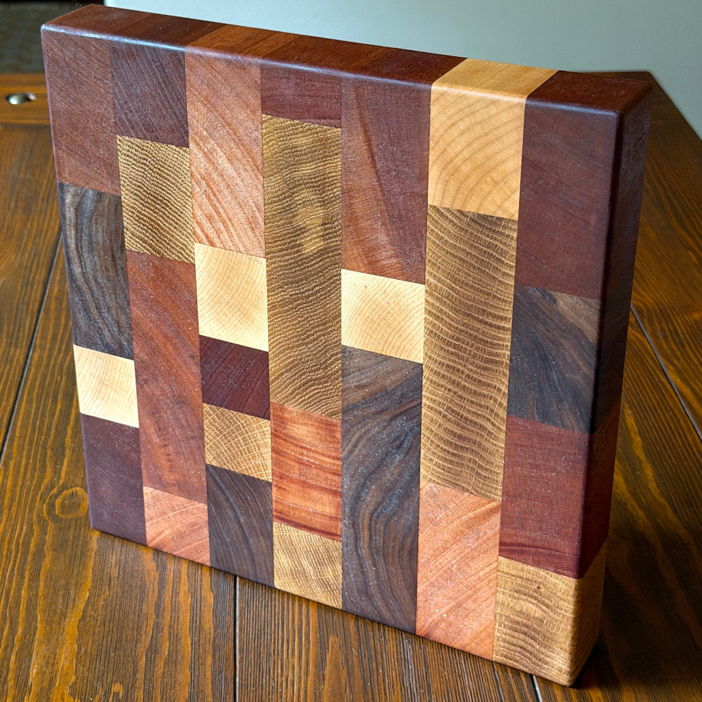 A rectangular wooden cutting board with a geometric mosaic pattern of various wood grain blocks in shades of brown, beige, and reddish wood, standing on a wooden table.