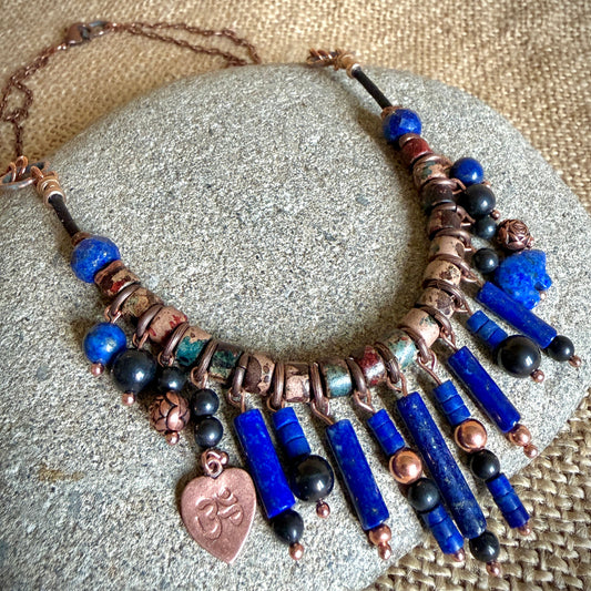 A bohemian-style statement necklace featuring lapis lazuli blue beads, shungite beads, copper spacers, and a dangling copper heart charm with a Sanskrit Om symbol, displayed on a gray stone over a burlap background.