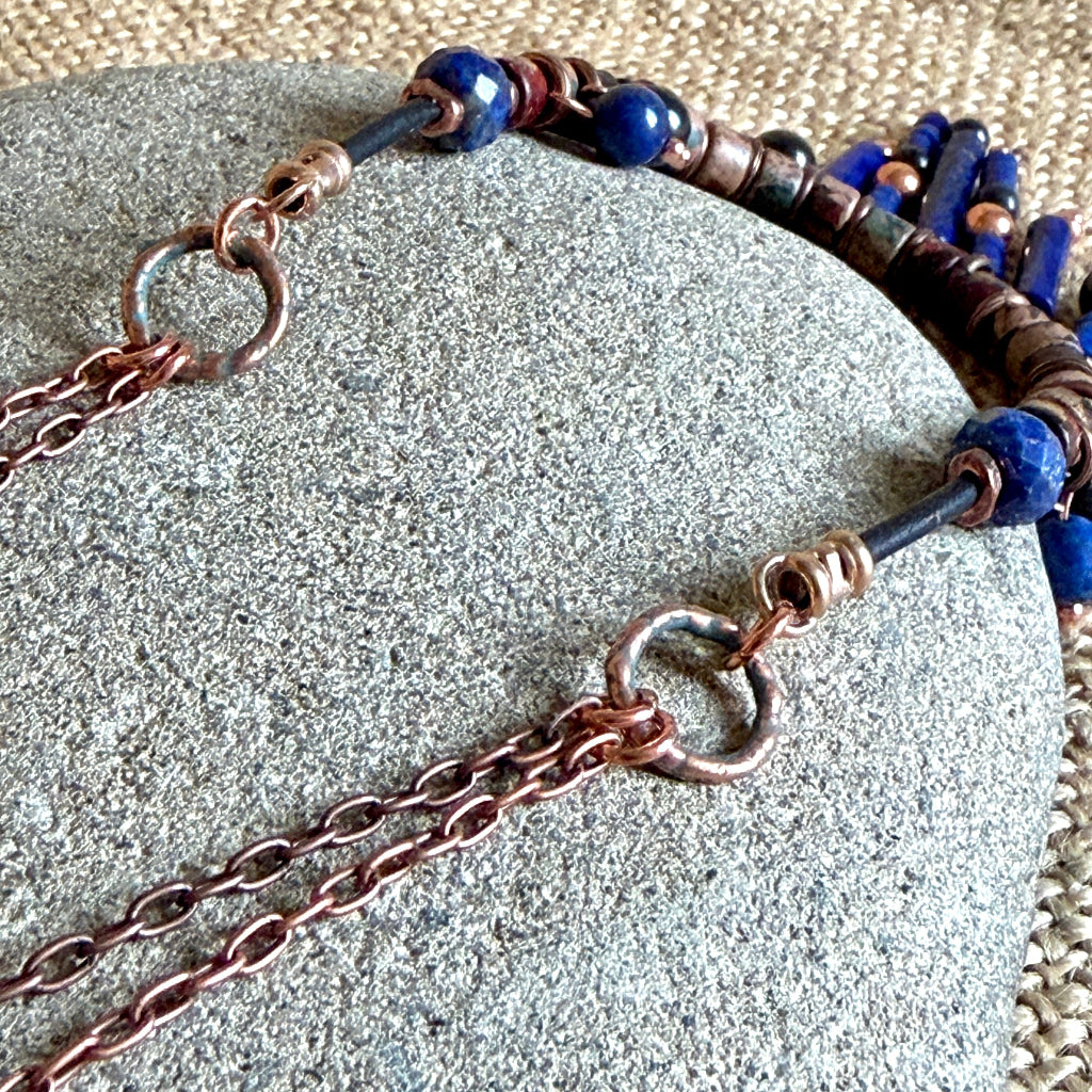 A close-up of a handmade beaded necklace draped over a gray stone. The necklace features dark blue lapis lazuli beads, copper metal spacers, and a beaded section.