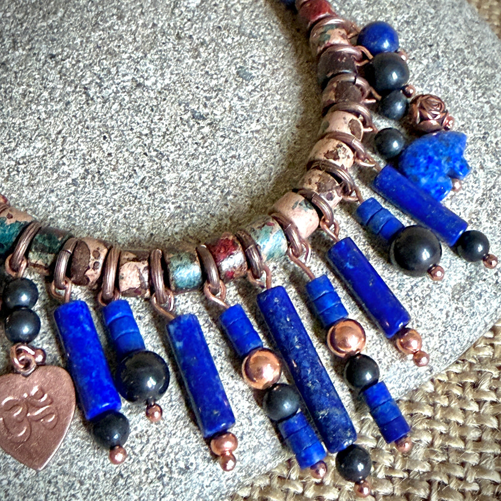 A bohemian-style beaded necklace featuring vibrant blue lapis lazuli tubes, shungite beads, ceramic spacers, and a small heart-shaped copper charm engraved with an Om symbol, displayed on a textured gray stone surface.