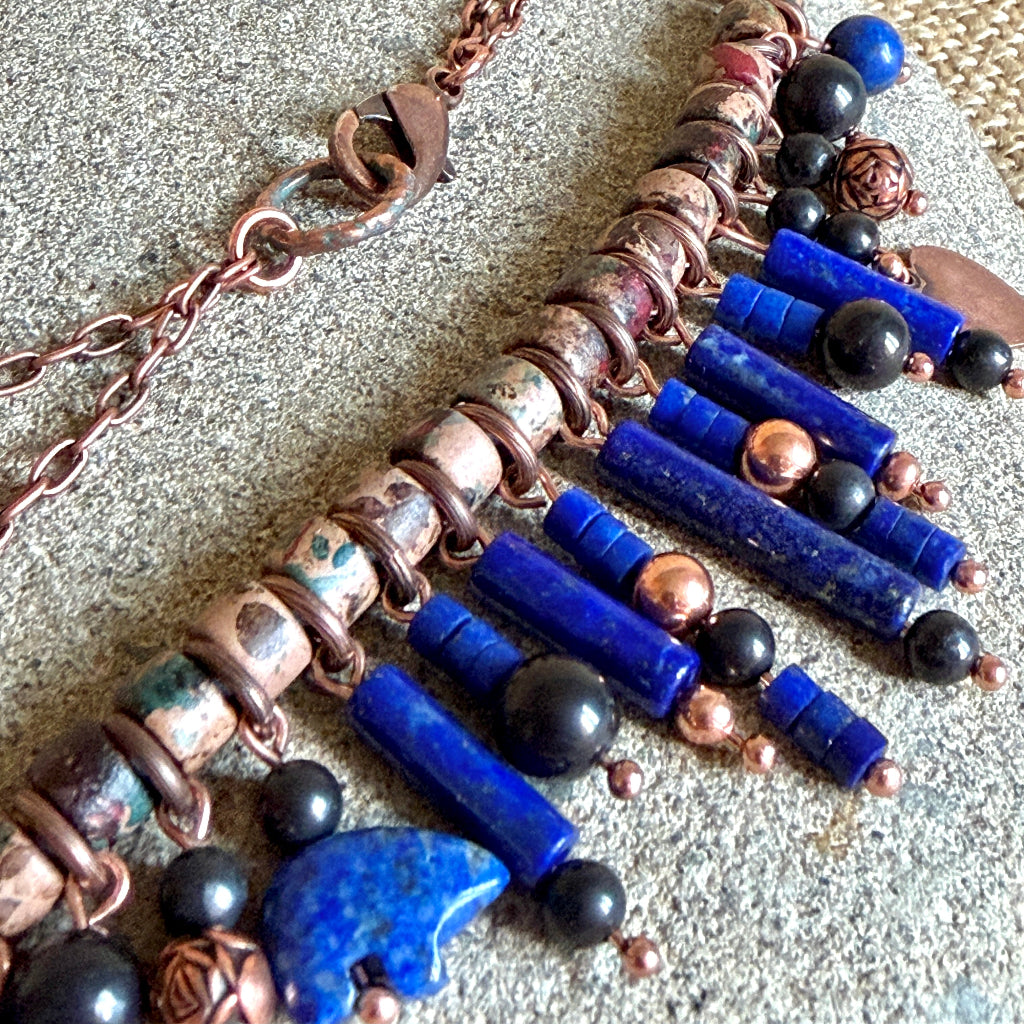 A close-up of a handmade statement necklace featuring vibrant blue lapis lazuli cylinder beads, shungite beads, patterned ceramic spacers, and a copper chain clasp, laid on a textured gray stone surface.